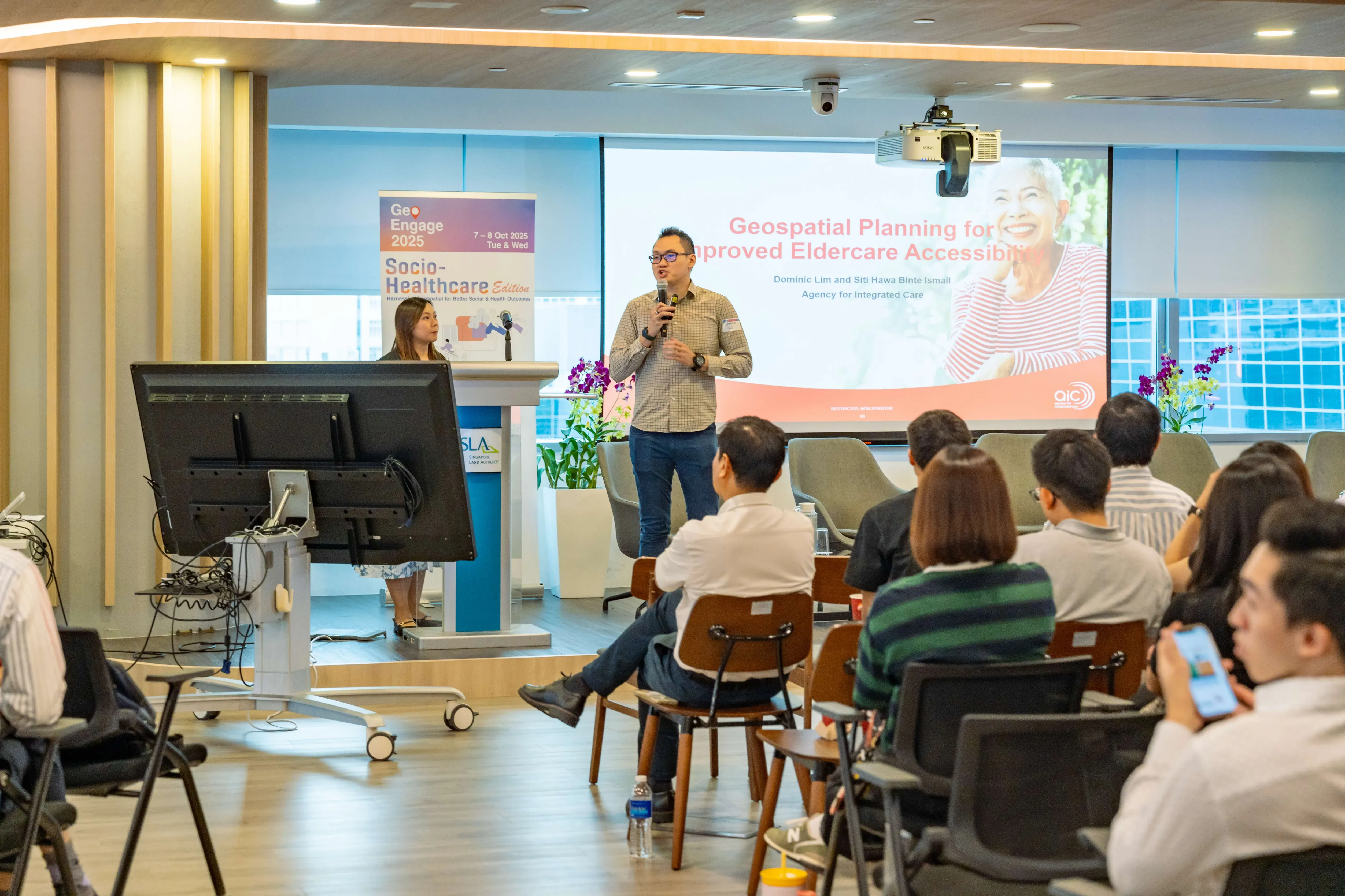 A man speaks at GeoEngage 2025 conference on Geospatial Planning for Eldercare Accessibility to a seated audience.